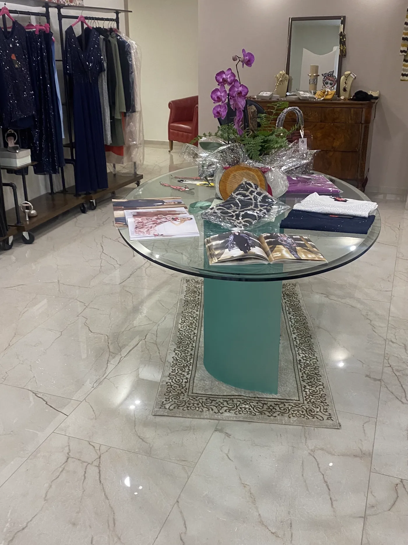 Interno de La Boutique della Moda: tavolo centrale in vetro con orchidee, abiti scuri esposti sul lato e mobile classico con specchio e accessori sul fondo